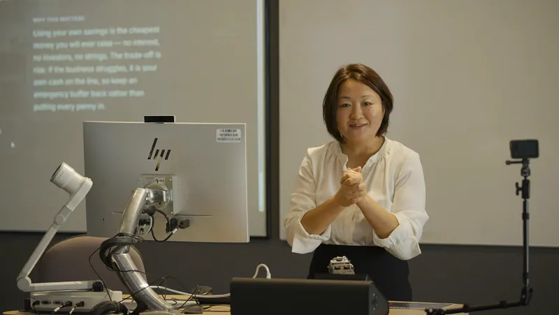 A presenter at a lectern beside a projected explainer on funding a business from your own savings.